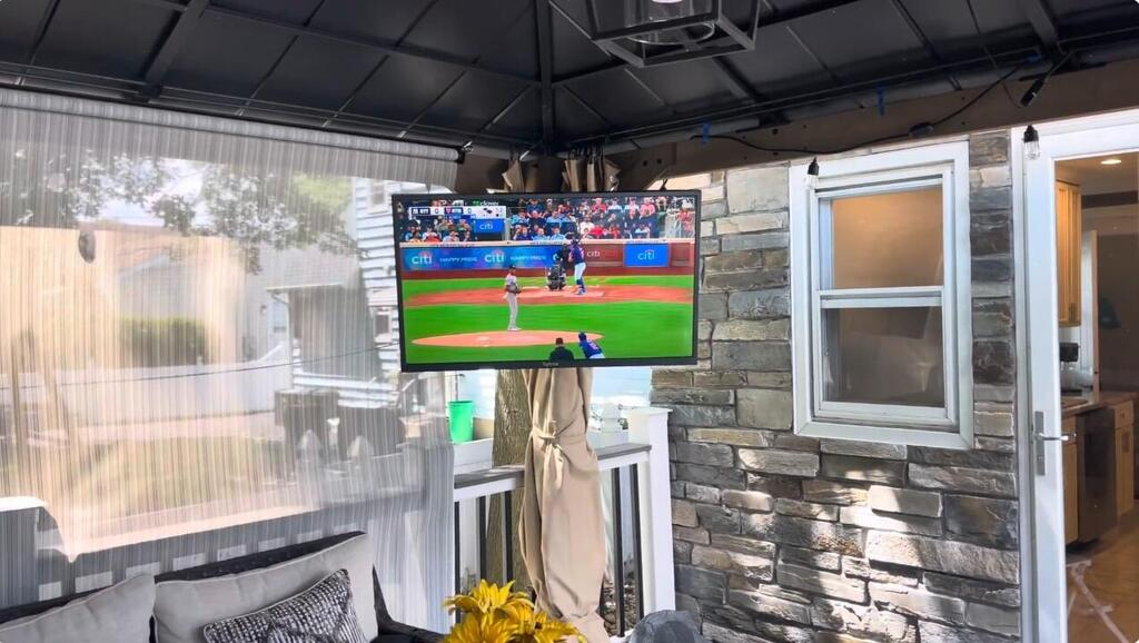 Outdoor living space with a TV displaying a sports game, surrounded by patio furniture and decor.