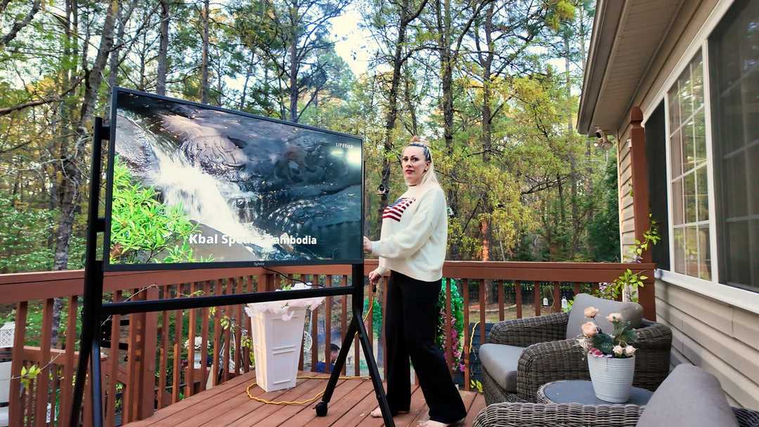 Woman standing on a deck with a large screen displaying nature scenery, surrounded by trees and outdoor furniture.